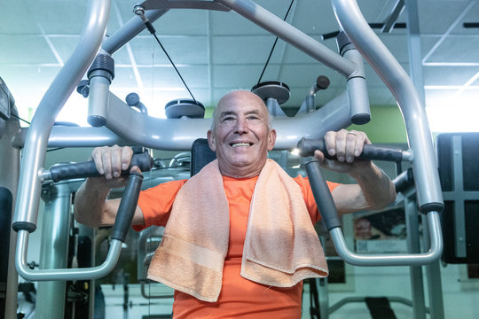 Uomo anziano con maglia arancione e asciugamano al collo  si allena in palestra nei macchinari, 