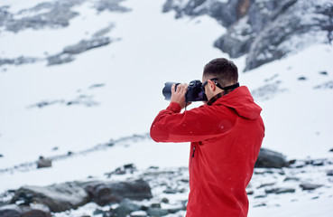 Back view of man taking photo with professional camera. Guy wearing red jacket while photographing winter nature. Concept of travelling, hiking and professional photography.