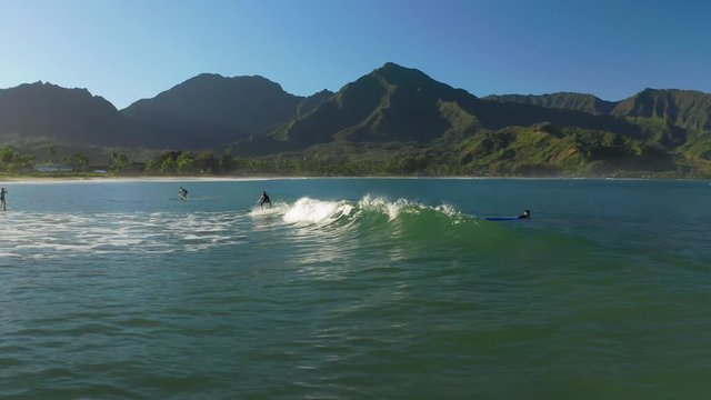 Drone footage. Watching the surfers riding ocean waves against the backdrop of picturesque coastal mountain range under clear blue sky. Hanalei, Hawaii. Travel destination. Aerial, 4K