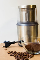 Coffee grinding at home. Electric metal coffee grinder, roasted coffee beans and ground coffee in a glass bowl on a beige table.