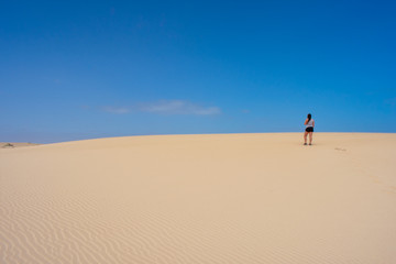 Fototapeta na wymiar Beautiful Young tanned, brunette woman, walking on sand dunes or desert against blue sky. Corralejo, Fuerteventura, Canary Islands, Spain 