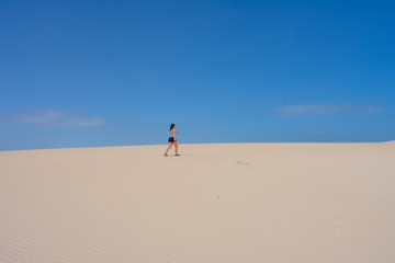 Fototapeta na wymiar Beautiful Young tanned, brunette woman, walking on sand dunes or desert against blue sky. Corralejo, Fuerteventura, Canary Islands, Spain 