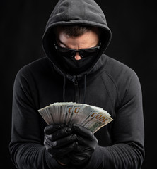 Man in black mask and hoody holding pack of dollars