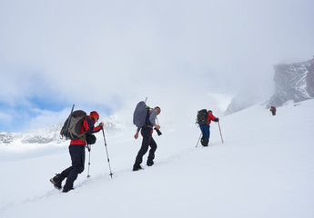 Group of brave tourists with backpacks and trekking sticks walking uphill in winter mountains,...
