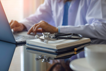 Female doctor in white lab coat typing on laptop computer with notebook and medical stethoscope on...