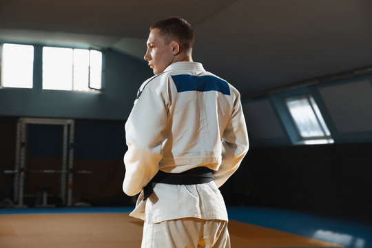 Young judo caucasian fighter in blue kimono with black belt posing confident in the gym, strong and healthy. Practicing martial arts fighting skills. Overcoming, reaching target, self building up.
