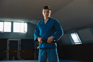 Young judo caucasian fighter in blue kimono with black belt posing confident in the gym, strong and healthy. Practicing martial arts fighting skills. Overcoming, reaching target, self building up. - obrazy, fototapety, plakaty