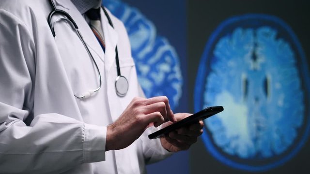 A medical doctor in a white lab coat and a stethoscope working on his mobile phone to look at a brain MRI scan image result, and typing in some information about his patient's health. Public Domain ba