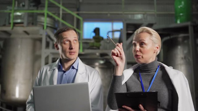 Medium shot of male engineer with laptop and female quality control expert with clipboard walking down factory and inspecting production processes