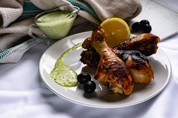 Toasted chicken drumsticks, with a brown Golden crust with fresh sauce with herbs decorated with lemon and black olives on a platter with a white textured napkin, towel