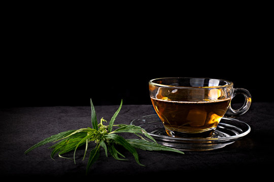 Cannabis tea in a clear glass cup with a green marijuana leaf and stem on wood table at the black background, Herbal and healthy drinks concept