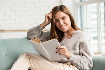  Pretty woman sitting on cozy sofa and reading book. 