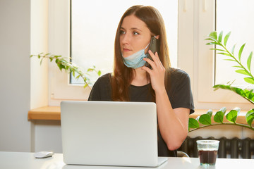 A woman in the removed medical face mask working remotely on her laptop during the quarantine to...