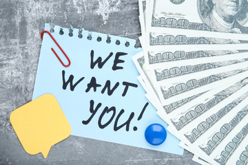 Text We Want You on paper with dollar banknotes on grey background
