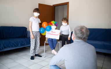 Children are congratulating their father in the time of pandemic. Happy Birthday is written in Italian.