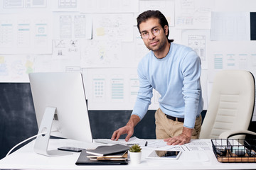 Portrait of confident middle-aged man with stubble standing at desk in office and using computer to...