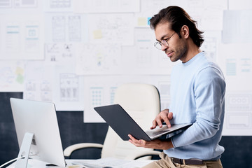 Serious modern middle-aged man in glasses using laptop while analyzing interfaces of web sites in UI design office