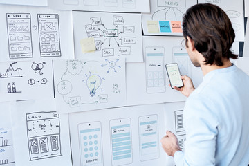 Rear view of brown-haired man testing new app on smartphone while working on interface design in...