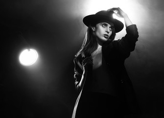 Beautiful braless actress girl, wearing an unbuttoned blazer and a hat, poses and dances in the warm light rays of scenic illuminators in the theatrical fog. Artistic, monochrome, noir design.