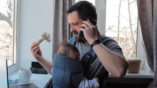 Busy Dad with baby in carrier at home talking on the phone.
