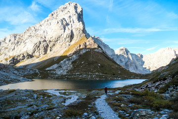 A woman in hiking outfit walking along the Wolayer Lake in Austrian Alps. There is massive, rocky mountain on the other side of the lake. New day beginning. Soft reflections in the lake. Happiness