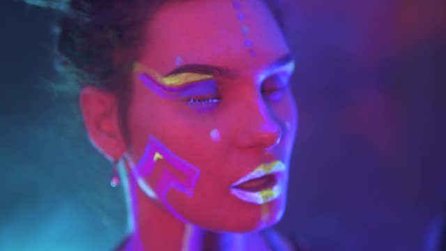 Making up a Girl with Dreadlocks in Neon UF Light. Model Girl with Fluorescent Creative Psychedelic MakeUp, Art Design of Female Disco Dancer Model in UV, Colorful Abstract Make-Up. Dancing Lady