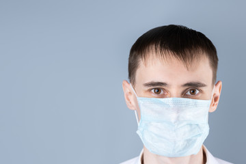 Man in a medical mask.  Close up of a handsome dark-haired, brown-eyed man in a white medical coat and medical mask  looking at camera , on a gray background. Chinese Coronavirus , Covid-19