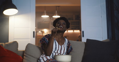 Happy young millennial African woman smiles watching TV soap opera series at home on the couch with snacks slow motion.