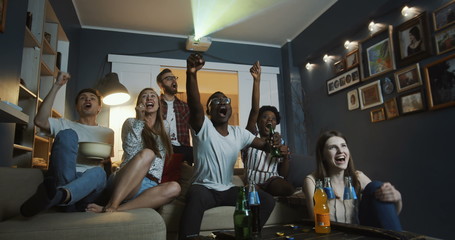 Excited young happy multiethnic friends get emotional and shout watching sports on TV together at...