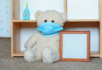 Quarantine, stay at home, no kindergarten: teddy bear in medical face mask sits with thermometer near children playhouse, holds poster, empty picture frame, place for inscription copy spaсe