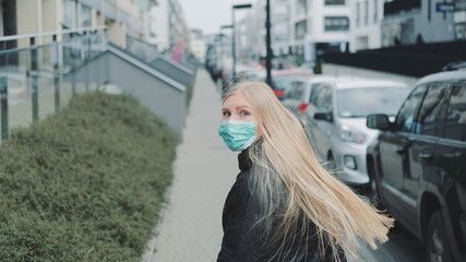 Young blonde woman in a medical mask escaping from somebody on the street. She is scared and concerned. Coronavirus pandemic concept.