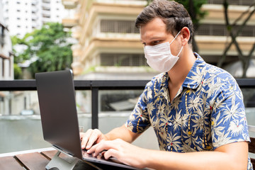 Young man with protective mask on his face working remotely with his laptop