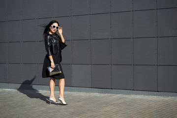 Street fashion look. Beautiful young woman in stylish sunglasses wearing elegant dress and fashionable black fringed leather jacket holding clutch in the hands.  Urban wall on background. Copy space.