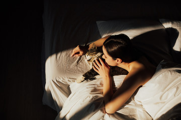 Fototapeta na wymiar Beautiful young woman with cute cat lying in bed at home. She tender hugging his pretty cat in sunny morning. Shadows on bed, copy space. 