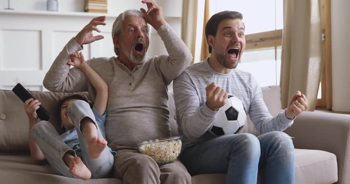 Overjoyed emotional three male generations family watching football soccer championship, spending fun time together at home. Happy elder man supporting favorite sport team with son and grandson.