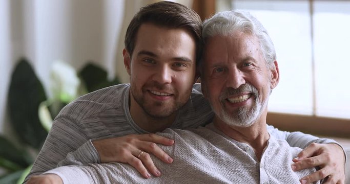 Handsome young man embracing shoulders of happy elderly mature grey haired father, looking at camera. Smiling two male generations family posing for portrait at home, showing love care devotion.