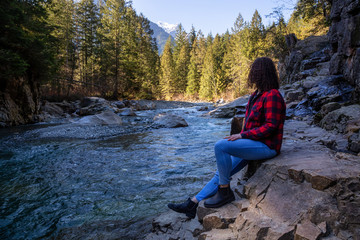 Naklejka na ściany i meble Girl Sitting by the river in the Canadian Mountain Landscape during a sunny winter day. Taken in Golden Ears Provincial Park, near Vancouver, British Columbia, Canada.