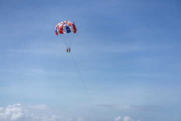 From a distance, two tourists are enjoying tandem parasailing while on holiday in Bali. Blue sky and white clouds appear.