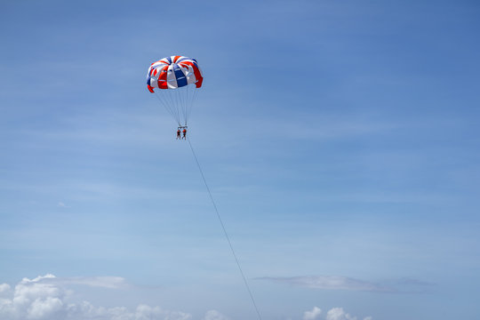 From a distance, two tourists are enjoying tandem parasailing while on holiday in Bali. Blue sky and white clouds appear.