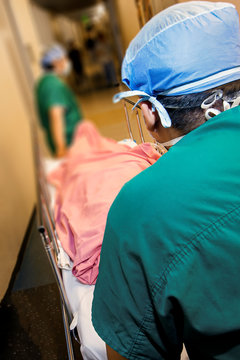 Image with perspective of following Medical Professionals in green scrubs pushing a gurney down hospital hallway with patient covered in pink blanket
