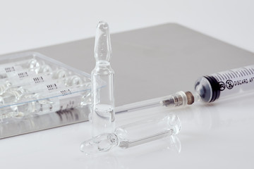 Upright ampoule with medicine next to ampoule and disposable syringe lying on white acrylic table,...