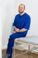 Portrait of a young male doctor. Man in blue uniform of doctor sits on massage table in massage room
