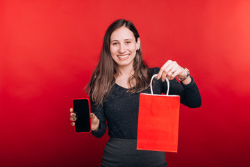 Fototapeta na wymiar Look, I bought this online. Happy young woman is smiling at the camera showing a shopping bag and a mobile phone on red background.