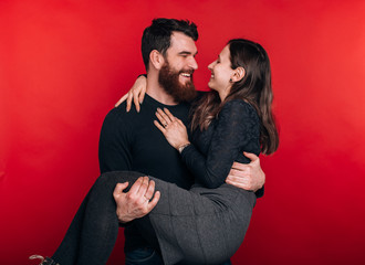 Happy bearded man si holding his woman on red background. Cheerful couple is enjoying eachother.