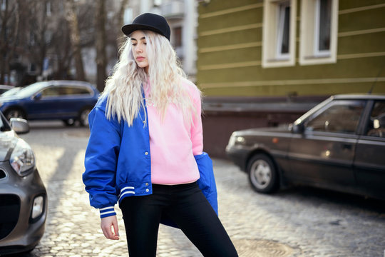 Female model with white blonde hair, posing on the street. Youth style fashion - black cap, blue baseball jacket and pink blouse. City background - buildings, cars on the street.