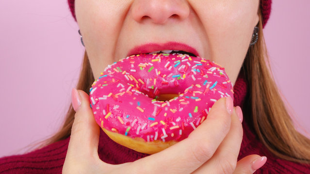 Beautiful adult female in sweater and hat biting delicious donut against pink background. Close up