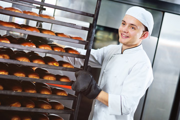 a young cute Baker in a white uniform smiles and puts an iron tray with bread and rolls on a shelf against the background of a bread factory or bakery.