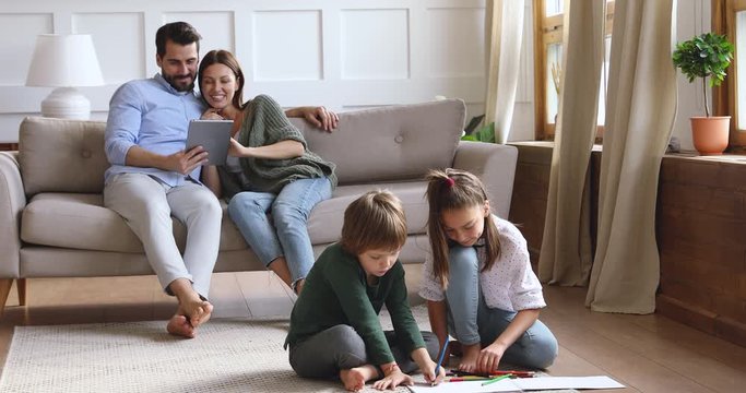 Happy young couple resting on sofa, watching funny movie online on tablet while small children siblings coloring drawing pictures in paper album, sitting together on floor carpet in living room.