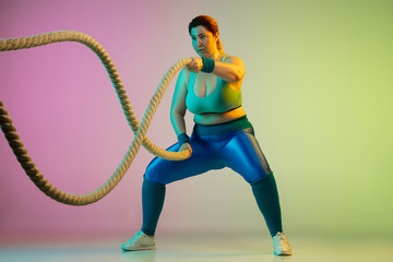 Young caucasian plus size female model's training on gradient purple green background in neon light. Doing workout exercises with ropes. Concept of sport, healthy lifestyle, body positive, equality.