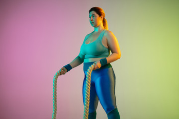 Young caucasian plus size female model's training on gradient purple green background in neon light. Doing workout exercises with ropes. Concept of sport, healthy lifestyle, body positive, equality.